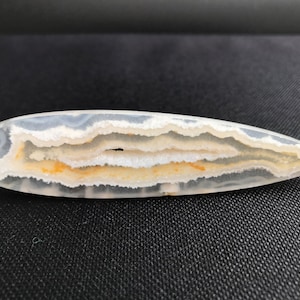 Könnte beinhalten: Ein polierter, länglicher Edelstein mit spitzem Ende. Der Stein weist Schichten aus weißen, grauen und gelb-orangen Farbtönen auf, die ein gebändertes Muster bilden. In der Mitte ist eine kleine Höhle zu sehen. Die Form erinnert an ein Blatt oder einen Tropfen.