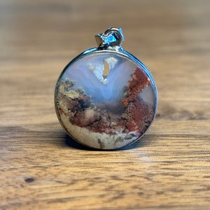 Può includere: Un ciondolo rotondo in argento con una pietra di agata bianca, marrone e arancione. La pietra presenta un motivo unico di vortici e linee.