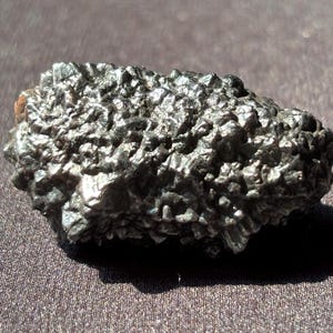 May include: A dark, textured rock with a rough, uneven surface. The rock is primarily dark gray with hints of brown and is illuminated by sunlight. The object is likely a mineral specimen or a unique geological formation.