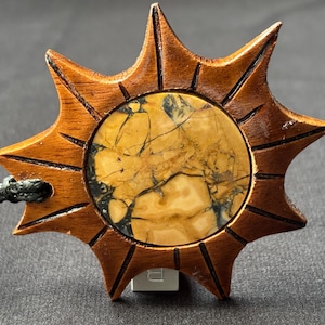 May include: A wooden pendant with a sunburst design. The pendant features a circular stone with a yellow and brown pattern, set within a star-shaped frame. A black cord is attached for wearing.