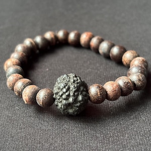 May include: A bracelet made of dark brown wooden beads with a central, textured, dark grey bead. The bracelet features round wooden beads, and the central bead has a unique, irregular surface. The bracelet is displayed on a dark background.