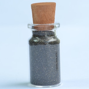 Puede incluir: Una pequeña botella de vidrio con un tapón de corcho llena de semillas de amapola negras.