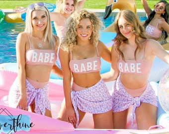 Op de afbeelding: Een groep mensen draagt bijpassende roze badkleding met het woord "BABE" op de tops. Ze bevinden zich in een zwembad met opblaasbare drijvers. De achtergrond toont een kleurrijke muurschildering met de woorden "Good Vibes".