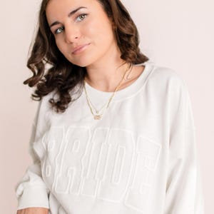 A woman with dark, wavy hair wearing an oversized white sweatshirt with the word "BRIDE" embossed across the front. She is accessorized with delicate gold necklaces and rings, posing softly against a neutral backdrop.