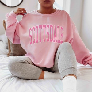 May include: A light pink sweatshirt with the word "SCOTTSDALE" in a contrasting pink block letter design. The sweatshirt is paired with gray sweatpants and white socks. The person is sitting on a white bed.