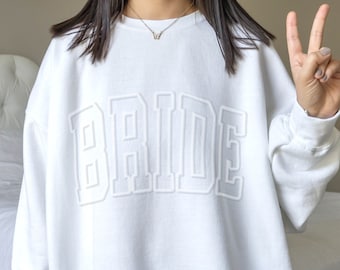 Op de afbeelding: Wit sweatshirt met het woord "BRIDE" in grote, reliëf, witte letters.