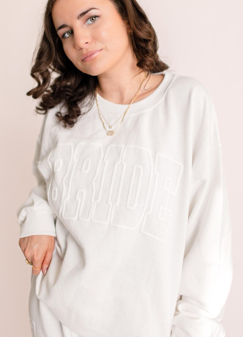 A woman with dark, wavy hair wearing an oversized white sweatshirt with the word "BRIDE" embossed across the front. She is accessorized with delicate gold necklaces and rings, posing softly against a neutral backdrop.