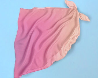 Op de afbeelding: Een roze en oranje ombre bandana met een geknoopte hoek. De bandana is gemaakt van een zachte, vloeiende stof en ligt plat op een lichtblauwe achtergrond.