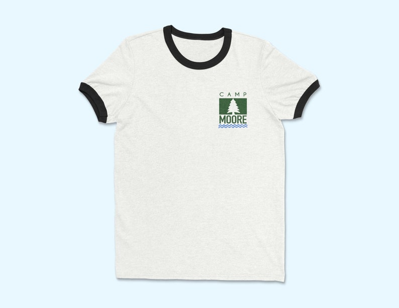 以下が含まれることがあります： 白地に黒のラインが入ったTシャツで、緑色の松の木のロゴがあり、白い枠の中に「Camp Moore」と書いてあります。