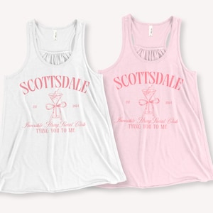 May include: Two tank tops with a pink graphic design that says "Scottsdale Est. 2024 Invisible String Social Club Tying You To Me". One tank top is white and the other is pink.