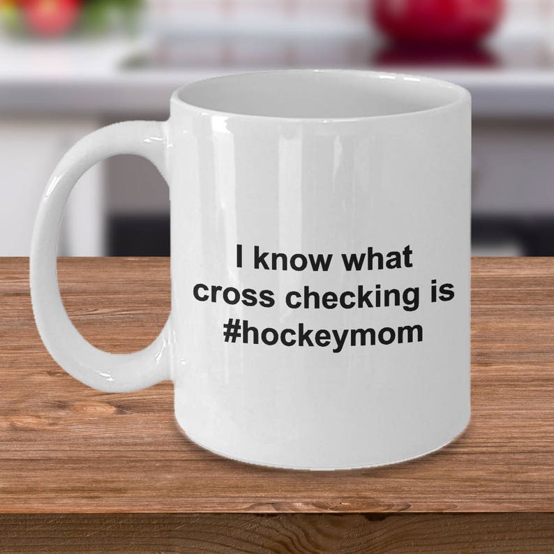 Może przedstawiać: Biały ceramiczny kubek do kawy z czarnym napisem "Wiem, co to jest cross-checking #hockeymom".