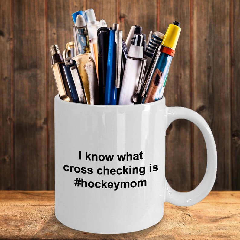 Może przedstawiać: Biały ceramiczny kubek do kawy z czarnym uchwytem i nadrukiem "I know what cross checking is #hockeymom" na przodzie. Kubek jest wypełniony r&oacute;żnymi długopisami i oł&oacute;wkami.