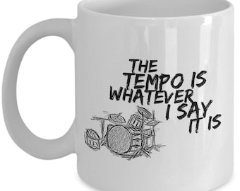 May include: White ceramic mug with a black sketch of a drum set and the text "THE TEMPO IS WHATEVER I SAY IT IS". The mug has a curved handle and is suitable for hot or cold beverages.
