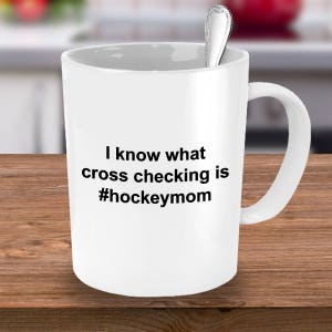 Może przedstawiać: Biały ceramiczny kubek do kawy z czarnym nadrukiem tekstu: "Wiem, co to jest cross-checking #hockeymom".
