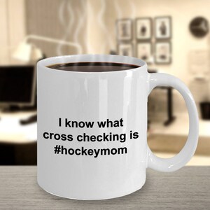 Może przedstawiać: Biały ceramiczny kubek do kawy z czarnym uchwytem. Kubek ma czarny napis "I know what cross checking is #hockeymom".