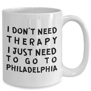 May include: White ceramic mug with the text "I DON'T NEED THERAPY I JUST NEED TO GO TO PHILADELPHIA" in black, bold letters. The mug has a curved handle and is designed for hot beverages.