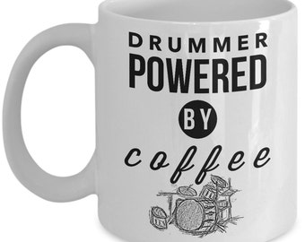 May include: White ceramic coffee mug with black text that reads "Drummer Powered by Coffee". A black circle with the word "By" is between the words "Powered" and "Coffee". A black and white illustration of a drum set is below the text.