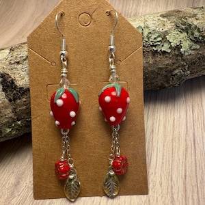 May include: A pair of dangle earrings featuring red strawberry beads with white polka dots and green tops. The earrings have silver-tone hooks and chains, with red flower and gold leaf charms.