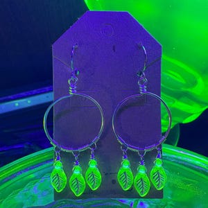 May include: A pair of silver hoop earrings with dangling neon green leaf charms. The earrings are displayed on a brown tag against a backdrop of green and purple hues, creating a vibrant and eye-catching aesthetic. The leaf charms have detailed vein patterns.