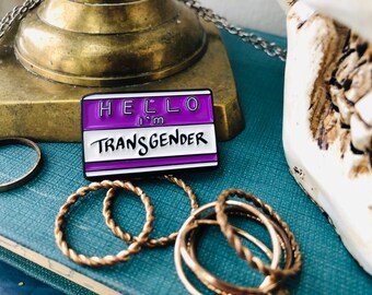 Può includere: Spilla rettangolare in smalto con la scritta "HELLO I'm TRANSGENDER" in bianco e nero su sfondo viola. La spilla è circondata da anelli color oro e poggia su una superficie blu.