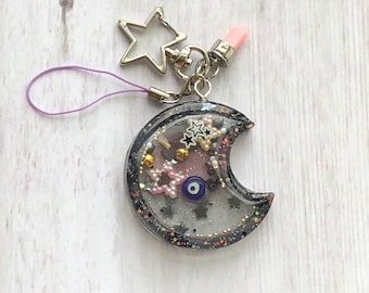 Può includere: Un ciondolo shaker a forma di mezzaluna con bordo glitterato nero, riempito con piccole paillettes a forma di stella, perline dorate e una perla a forma di occhio blu. Include una chiusura a stella argentata, un piccolo ciondolo rosa e un cordino viola.