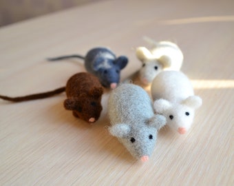 Può includere: Cinque giocattoli a forma di topo in feltro di diversi colori, tra cui grigio, marrone e bianco. I topi sono sdraiati su una superficie di legno chiaro.