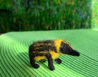 May include: A handmade felted animal figurine of a badger. The badger has a black head and legs, with a yellow and black striped body. The figurine is set against a green background.