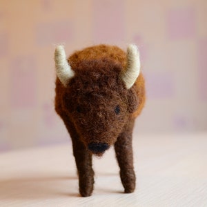 May include: A brown and white felt bison figurine with a fluffy texture. The bison is standing on a white surface.