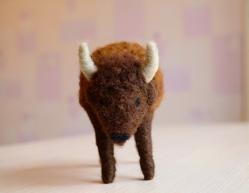 May include: A brown and white felt bison figurine with a fluffy texture. The bison is standing on a white surface.