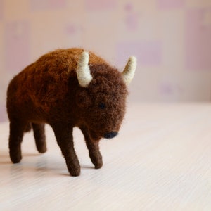 May include: A brown and white felt bison figurine with a fluffy texture. The bison is standing on a light wood surface.