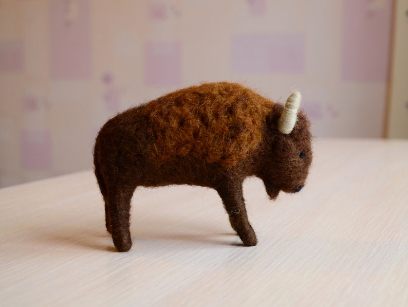 May include: A brown and white felt bison figurine. The bison is standing on a light wood surface.