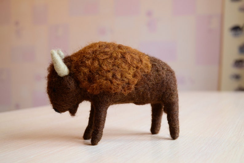 May include: A brown and white felt bison figurine. The bison is standing on a white surface.