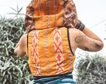 Peut inclure: Un gilet sans manches orange et marron à motifs avec une capuche assortie. Le gilet présente un motif géométrique rouge et blanc. La capuche et le gilet sont portés sur un haut turquoise et un short en jean blanc.