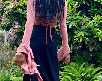 May include: A person wearing a pink long-sleeved shirt with a black skirt and braided hair. The person is walking away from the camera in a garden setting.