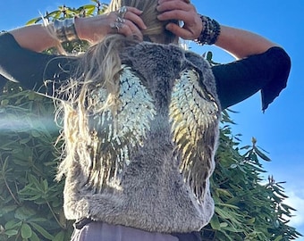May include: A woman wearing a brown faux fur vest with gold sequined angel wings on the back. She is wearing a purple ruffled top, gray jeans, and floral patterned boots.