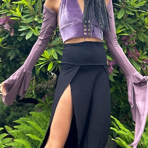 Puede incluir: Una mujer lleva una falda negra con una abertura alta y un top de terciopelo morado con tirantes. La falda es larga y fluida, y el top tiene un cierre de encaje en la parte delantera. La mujer está de pie frente a un arbusto verde.