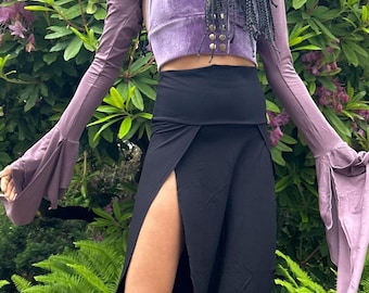 May include: A woman wearing a black skirt with a high slit and a purple velvet halter top. The skirt is long and flowy, and the top has a lace-up front. The woman is standing in front of a green bush.