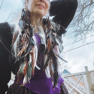 May include: A woman with long white hair wearing a black jacket, a purple top, and a black lace skirt. She is wearing long feather earrings with brown, green, and black feathers.