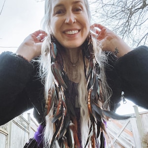 May include: A woman with long, white hair wearing a black jacket and a purple and black dress with long feather earrings. The earrings are made of black, brown, and teal feathers.