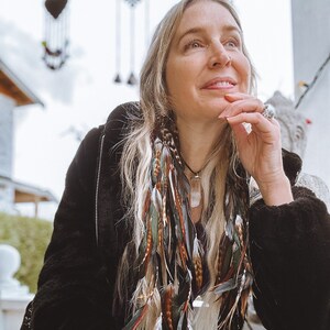 May include: A woman with long blonde hair wearing a black jacket with a zipper and a feather hair extension. The hair extension is made of brown, white, and green feathers. She is wearing a necklace with a large crystal pendant.