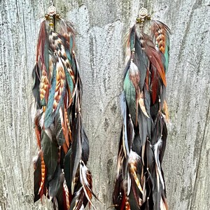 May include: A pair of long feather earrings with brown, black, green, and white feathers. The earrings have a beaded tassel and a metal charm at the top.