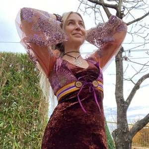 May include: A brown velvet mini dress with a purple and gold belt and a purple lace overlay. The dress has long sleeves with a purple and gold lace pattern. The model is wearing a silver necklace with a star pendant.
