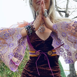 May include: A woman wearing a brown velvet dress with a purple and gold belt, a silver necklace with a pendant, and multiple bracelets on her wrists. She is wearing a sheer lace overlay with purple embroidery.
