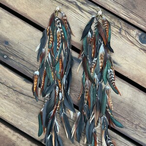 A pair of long, dangling earrings with feathers in shades of brown, teal, and black. The earrings are made with a variety of feathers some with a sheen.
