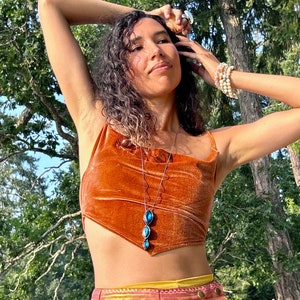 Może przedstawiać: Kobieta w koralowym aksamitnym topie na ramiączkach z niebieskim naszyjnikiem z kamieni szlachetnych i wielokolorową spódnicą ze złotym paskiem.