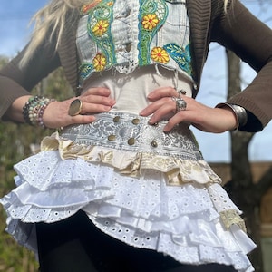 May include: A cropped light-wash denim vest with green and yellow floral embroidery, worn over a white top and a brown cardigan. The outfit includes a layered white eyelet lace skirt with a metallic silver patterned waistband and black leggings.