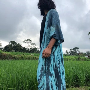 Peut inclure: Un long kimono ouvert sur le devant arborant un motif tie-dye aux nuances de turquoise vif et de noir charbon profond. Le v&ecirc;tement pr&eacute;sente des manches larges et amples ainsi qu'une silhouette fluide tombant jusqu'au sol.