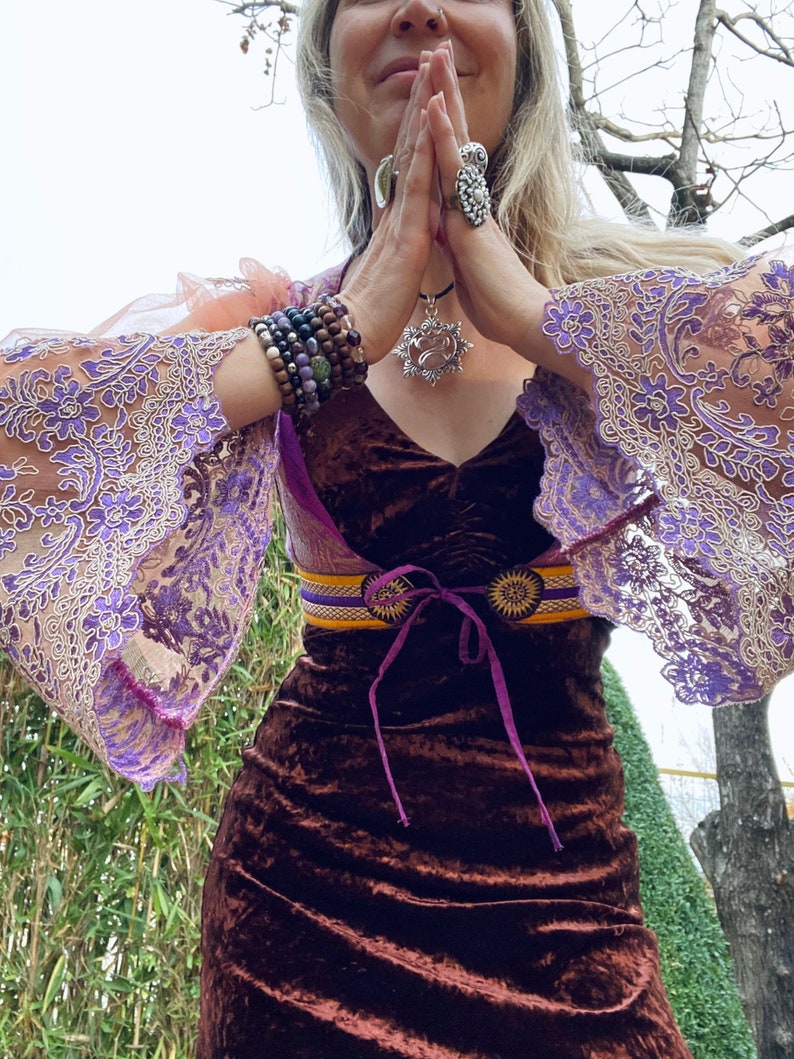 May include: A woman wearing a brown velvet dress with a purple and gold belt, a silver necklace with a pendant, and multiple bracelets on her wrists. She is wearing a sheer lace overlay with purple embroidery.