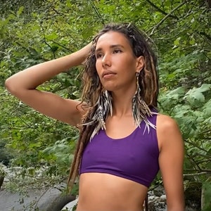 May include: A woman with long brown dreadlocks wearing a purple sports bra and feather earrings. She is standing in a forest setting.