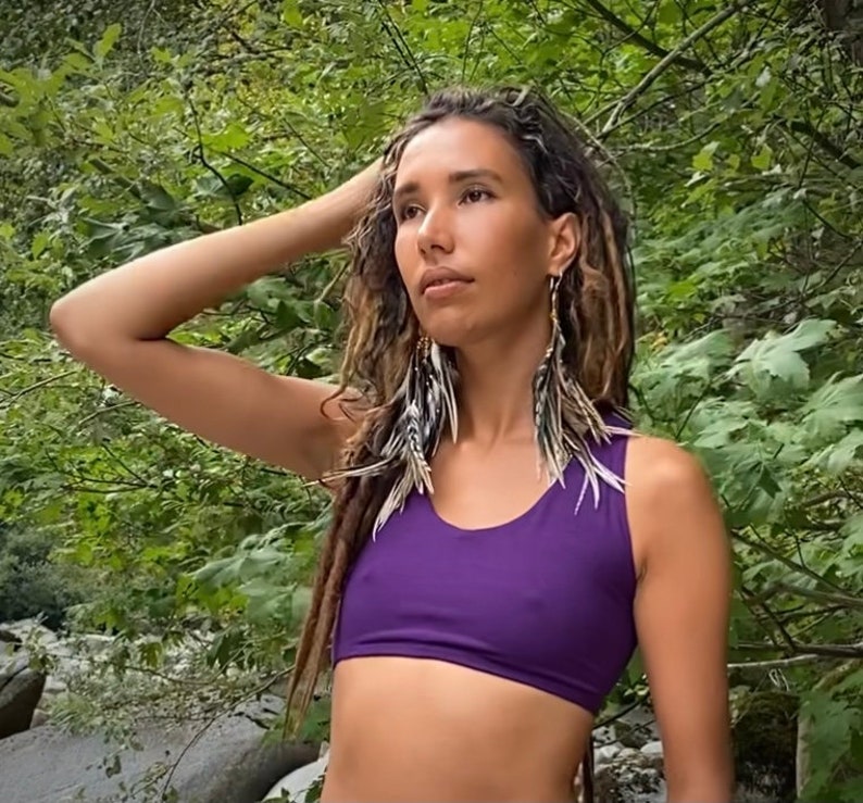 May include: A woman with long brown dreadlocks wearing a purple sports bra and feather earrings. She is standing in a forest setting.